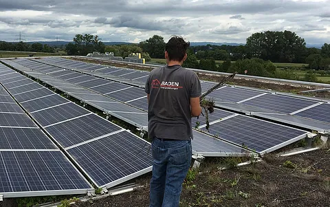 Mann in grauem T-Shirt steht auf einem Solarpanel-Dach und entfernt Unkraut zwischen den Solarmodulen.