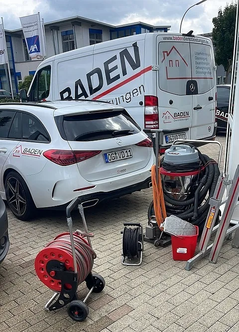 Zwei Fahrzeuge mit dem Schriftzug 'BADEN', ein weißer Mercedes und ein SUV, stehen auf einem Parkplatz. Neben den Fahrzeugen sind Werkzeuge und Schläuche für Reinigungsarbeiten sichtbar.