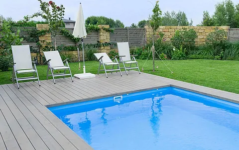 Ein moderner Garten mit einem blauen Pool, umgeben von Rasen und Pflanzen. Vier Liegestühle stehen am Poolrand, ein Sonnenschirm ist aufgestellt.