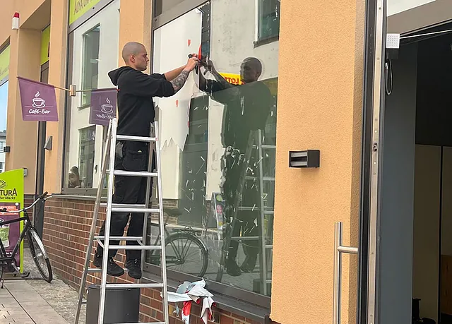 Ein Handwerker steht auf einer Leiter und bringt eine Folie an einem Schaufenster an. Im Hintergrund sind Fahrräder und ein Café-Schild sichtbar.