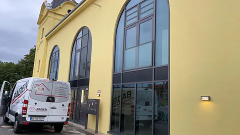 Gelbes Gebäude mit großen Fenstern und einem weißen Lieferwagen davor, der das Logo "Baden Dienstleistungen" trägt.