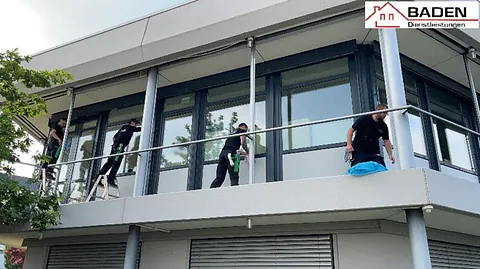 Drei Reinigungskräfte putzen Fenster an einem modernen Gebäude. Eine Person steht auf einer Leiter, während die anderen am Geländer arbeiten.