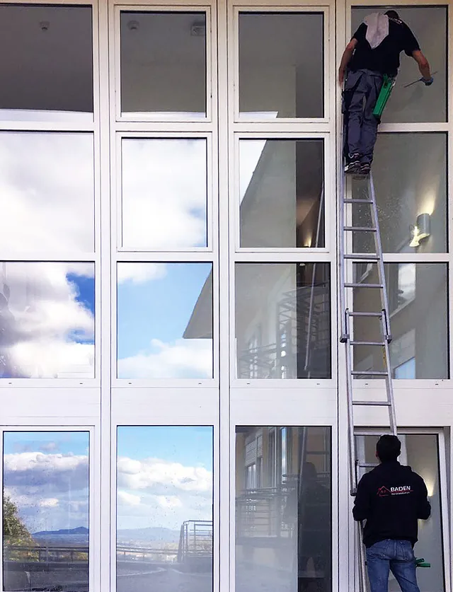 Fensterputzer auf einer Leiter reinigt große Fensterfront eines modernen Gebäudes, im Hintergrund Wolken und Landschaft sichtbar.