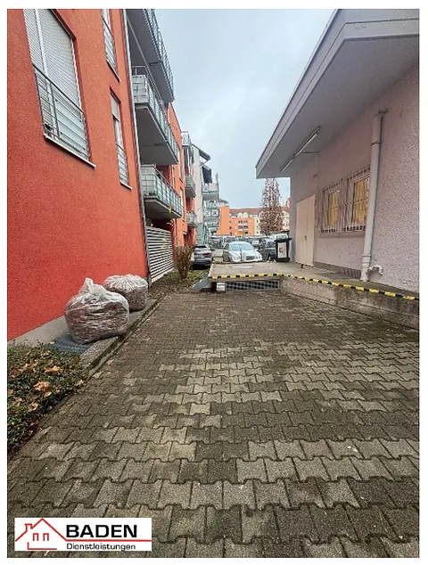 Blick auf einen gepflasterten Weg zwischen zwei Gebäuden, mit einem parkenden Auto und zwei großen Säcken auf der linken Seite. Im Vordergrund ist eine Absperrung sichtbar.
