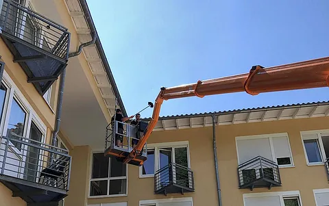 Arbeiter in einem Hubsteiger, der an einem Dachrand arbeitet, um Reinigungsarbeiten durchzuführen. Helle Fassade und blauer Himmel im Hintergrund.