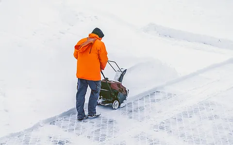 Mann in orangefarbener Jacke schiebt eine Schneefräse auf einem verschneiten Weg.