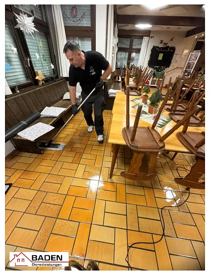 Ein Mitarbeiter reinigt den Boden eines Gasthauses mit einem Wischgerät. Tische und Stühle sind auf die Seite gestellt, um den Boden zu säubern.