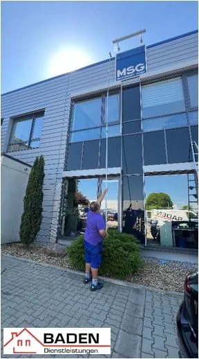 Ein Mann in lila Shirt und blauen Shorts steht vor einem modernen Gebäude und erreicht mit einer langen Stange ein Schild mit der Aufschrift 'MSG'. Im Vordergrund ist ein grüner Busch zu sehen.