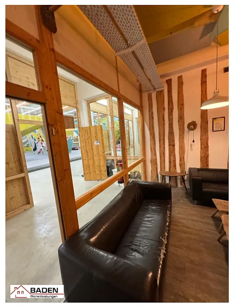 Innenansicht eines modernen Raumes mit Holzverkleidung, schwarzem Sofa und großen Fenstern, die auf einen hellen Bereich blicken.