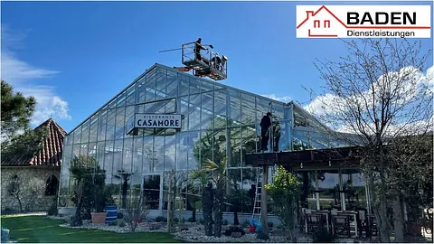 Glasgarten des Restaurants Casamore mit Handwerkern auf einer Hebebühne, die an der Fassade arbeiten. Logo von Baden Dienstleistungen oben rechts.