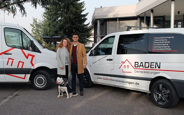 Paar steht neben zwei Fahrzeugen der Firma BADEN Dienstleistungen, mit einem Hund. Die Fahrzeuge sind mit dem Logo und der Website der Firma beschriftet.