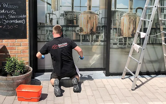 Mann in schwarzem T-Shirt mit Firmenlogo reinigt eine Glasfront vor einer Brauerei. Im Hintergrund sind große Braukessel sichtbar.