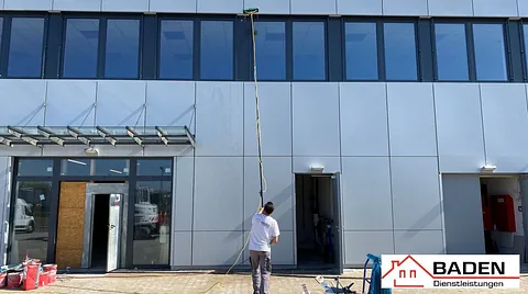 Ein Arbeiter reinigt mit einer langen Stange die Fenster eines modernen Gebäudes. Im Vordergrund sind Werkzeuge und Materialien sichtbar.