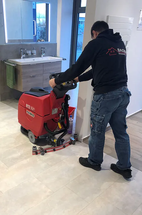 Ein Techniker in einem schwarzen Sweatshirt reinigt den Boden mit einer roten Reinigungsmaschine in einem modernen Badezimmer.