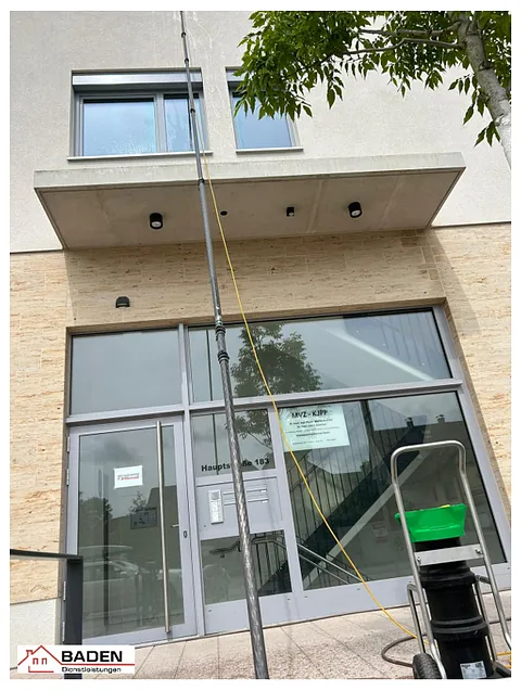 Fassade eines modernen Gebäudes mit großen Fenstern und einem Eingang. Ein Reinigungsgestell steht im Vordergrund, neben einer langen Stange.