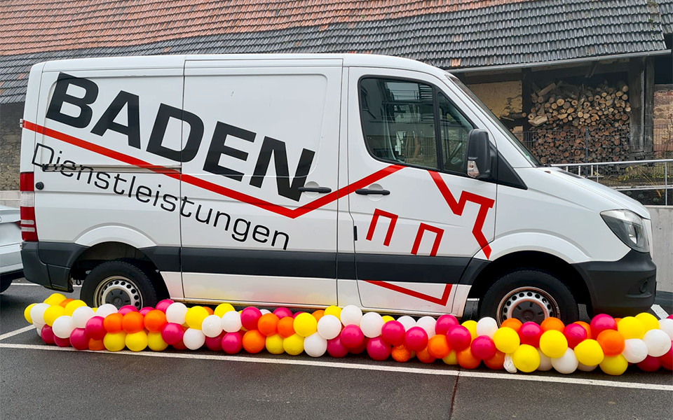 Weißer Lieferwagen mit der Aufschrift 'BADEN Dienstleistungen' und bunten Luftballons in Rot, Gelb und Weiß am Boden.